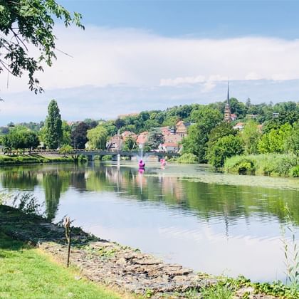 Rivière calme avec des berges vertes et des arbres en Franche-Comté. Un clocher d'église et des bâtiments de village sont visibles de l'autre côté de l'eau.