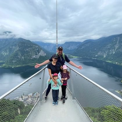 Famille de quatre personnes sur plateforme d'observation métallique surplombant le lac de Hallstatt et les montagnes du Salzkammergut autrichien.