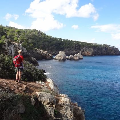 Randonneur avec sac à dos rouge sur falaise rocheuse de Majorque, contemplant les eaux turquoise de la Méditerranée et falaises calcaires.