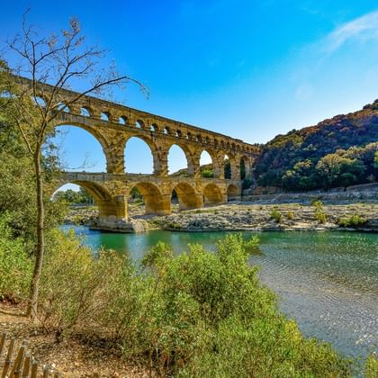 Ancien aqueduc romain du Pont du Gard avec trois niveaux d'arches enjambant le Gardon, entouré de collines vertes et végétation.
