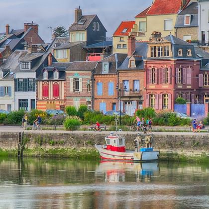 Rangée de maisons colorées à Saint-Valéry avec deux petits bateaux amarrés le long d'un quai en pierre. Des gens se promènent sur le front de mer.