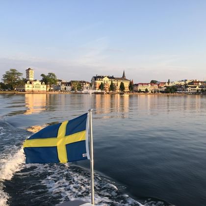 Drapeau suédois flottant sur un bateau avec le front de mer de Stockholm en arrière-plan. Bâtiments historiques et fontaine visibles le long du rivage.
