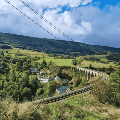 Viaduc ferroviaire en pierre traversant la campagne verdoyante du chemin Stevenson. Collines et forêts sous un ciel bleu avec nuages.