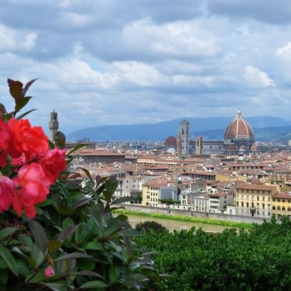 Fleurs rouges au premier plan avec le panorama de Florence derrière, montrant le dôme en terre cuite du Duomo et les clochers sous un ciel nuageux.