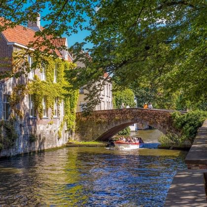 Canal pittoresque à Bruges avec bâtiments médiévaux couverts de lierre, pont de pierre et bateau touristique. Promenade bordée d'arbres.