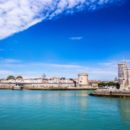 Vue du port de La Rochelle avec eau turquoise, tours médiévales en pierre à droite et bâtiments blancs avec clocher sous ciel bleu.