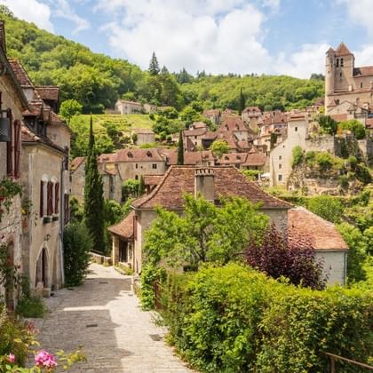 Rue pavée à Saint-Cirq-Lapopie avec maisons médiévales et église sur la colline. Verdure luxuriante autour des bâtiments aux toits de tuiles.