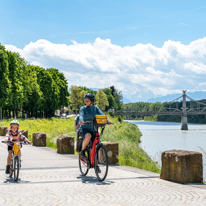 Adulte et enfant à vélo sur chemin riverain à Salzbourg avec la Salzach, pont et montagnes en arrière-plan sous ciel bleu nuageux.