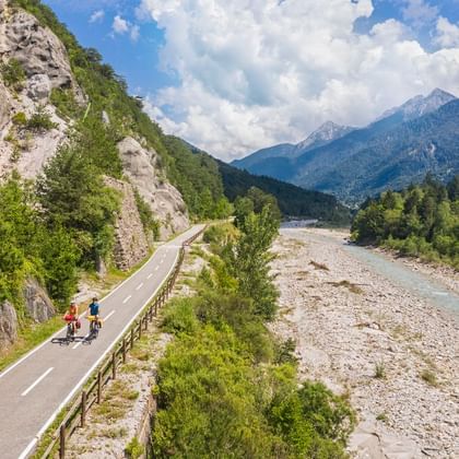 Deux cyclistes sur la piste cyclable Val Canale le long d'un lit de rivière rocheux, avec des falaises calcaires et des montagnes boisées.
