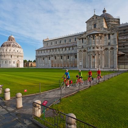 Quatre cyclistes sur un chemin près de la pelouse verte de la place de la cathédrale de Pise, avec la cathédrale en marbre blanc et le baptistère rond.