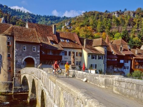 Cyclistes traversant le pont de pierre médiéval sur le Doubs à Saint-Ursanne, avec bâtiments historiques et collines boisées.