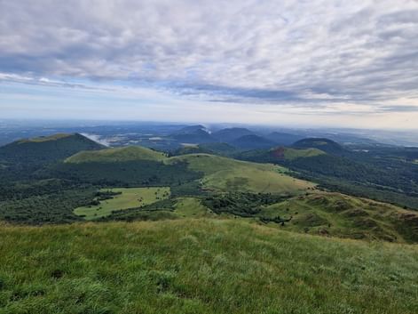 Vue étendue depuis le Puy de Dôme montrant des collines volcaniques vertes et des montagnes s'étendant à l'horizon sous un ciel nuageux.