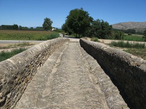 Ancien pont en pierre aux murs patinés traversant le paysage rural de la Costa Brava avec champs, arbres et collines sous ciel bleu.