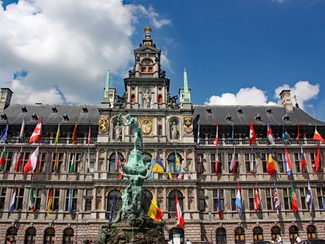 Hôtel de ville historique d'Anvers avec façade Renaissance ornée, tour d'horloge centrale et statue de bronze au premier plan. Drapeaux internationaux colorés bordent le bâtiment.