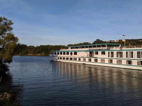 Bateau de croisière MS Switzerland naviguant sur un fleuve calme avec des arbres verts sur la rive gauche et des bâtiments visibles au loin.