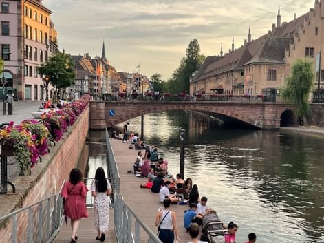 Personnes se détendant au bord d'un canal à Strasbourg. Un pont en pierre traverse l'eau, bâtiments historiques et clochers en arrière-plan.