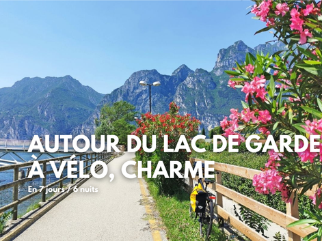 Piste cyclable au bord du lac de Garde avec lauriers-roses roses, montagnes en arrière-plan et vélo avec sacoches jaunes.