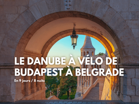 Vue à travers une arche en pierre avec lanterne suspendue, montrant un clocher et des arbres au coucher du soleil. Texte : Le Danube à vélo de Budapest à Belgrade.