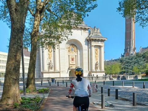 Cycliste s'approchant de la Porte de Paris, arc de triomphe blanc orné en Flandre française, avec arbres bordant la place et beffroi visible.