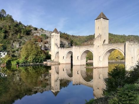 Pont médiéval Valentré avec trois tours et six arches enjambant le Lot, parfaitement reflété dans l'eau calme, entouré de collines vertes.