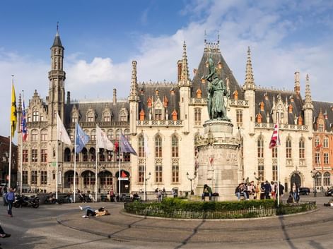 Palais Provincial de Bruges avec architecture gothique et flèches ornées. Un monument se dresse au centre de la place, entouré de touristes.