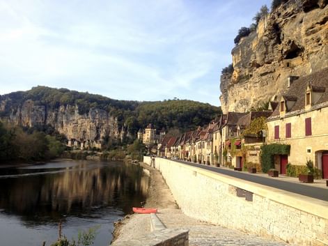 Maisons en pierre avec volets rouges le long d'une route au bord de la rivière en Périgord Noir, adossées à des falaises calcaires spectaculaires.