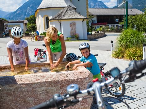 Mère et deux enfants avec casques font une pause à une fontaine en pierre pendant un tour à vélo, vélos à proximité et église alpine en arrière-plan.