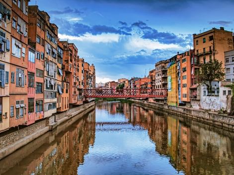 Des bâtiments colorés bordent les deux rives de la rivière Onyar à Gérone, avec un pont rouge traversant l'eau. Le ciel bleu avec des nuages blancs se reflète dans l'eau calme.