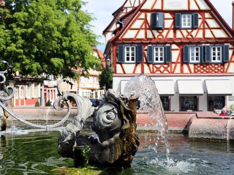 Fontaine ornée avec jet d'eau devant une maison à colombages rouge et blanc avec volets bleus dans une ville de la vallée du Rhin.
