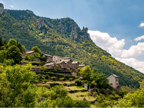 Village traditionnel en pierre sur une colline des Cévennes avec des champs en terrasses, entouré de forêts vertes et de pics rocheux.