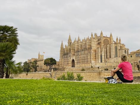 Cycliste en maillot rose assise sur l'herbe avec casque de vélo à côté, face à la cathédrale gothique de Palma à Majorque avec palmiers.