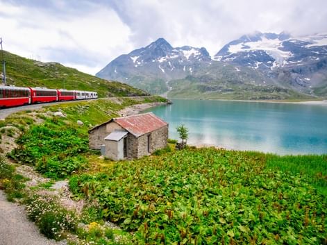 Train rouge Bernina Express longeant un lac alpin turquoise avec montagnes enneigées. Cabane en pierre et végétation verte au premier plan.