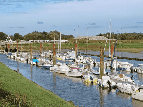 Petit port de plaisance avec voiliers blancs amarrés le long de quais en bois. Berges herbeuses encadrent le chenal, caravanes et dunes en fond.