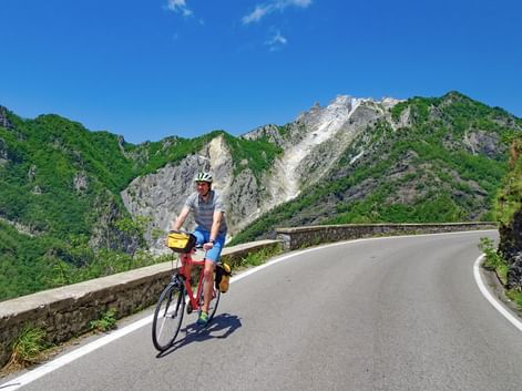 Cycliste avec casque et sacoches roulant sur route de montagne sinueuse avec garde-fou en pierre, entouré de montagnes boisées sous ciel bleu.