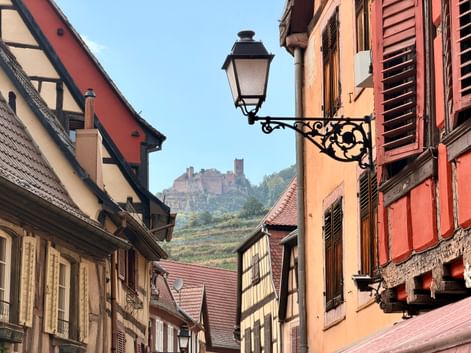 Maisons à colombages traditionnelles sur la Route des Vins d'Alsace avec lampadaire orné et château sur colline avec vignobles en arrière-plan.