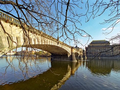 Pont en pierre avec arches enjambant la Vltava à Prague, avec le théâtre national visible sur la rive droite sous des arbres nus d'hiver.