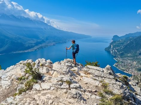 Randonneur avec sac à dos et bâtons sur sommet rocheux de Punta Larici, vue sur eaux bleues du lac de Garde entouré de montagnes.