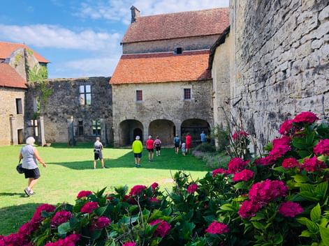 Cyclistes explorant une cour médiévale en pierre en Franche-Comté avec des passages voûtés et des fleurs roses au premier plan sous un ciel bleu.