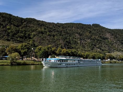 Bateau de croisière MS Switzerland naviguant sur eau verte calme avec collines boisées et petit village en arrière-plan sous ciel bleu.
