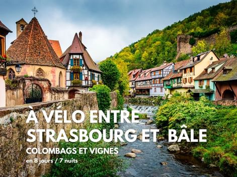 Maisons à colombages colorées bordent un ruisseau dans un village alsacien. Une chapelle au toit pointu se dresse à gauche, collines boisées derrière.