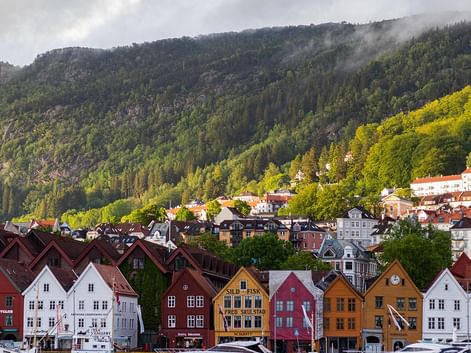 Rangée de maisons en bois colorées traditionnelles au bord de l'eau à Bergen, Norvège, avec montagnes boisées et nuages bas en arrière-plan.
