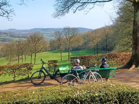 Deux cyclistes assis sur un banc vert surplombant les collines verdoyantes de Flandre française. Deux vélos sont garés à côté du banc sous un grand arbre.