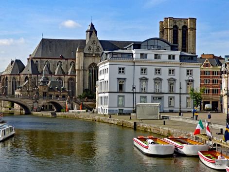 Front de mer historique de Gand montrant une cathédrale gothique avec haute tour, des bâtiments classiques blancs et des bateaux touristiques amarrés le long du canal.