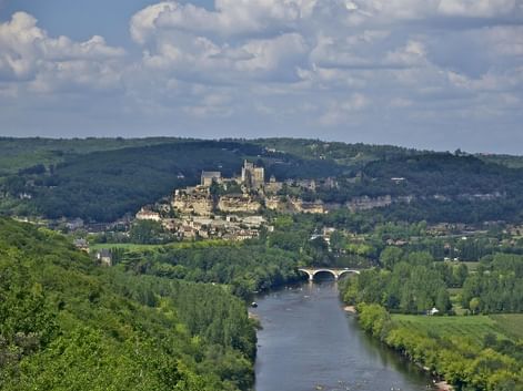 Château de Beynac perché sur une colline surplombant la Dordogne. Un pont de pierre traverse la rivière, entouré de forêts et collines vertes.