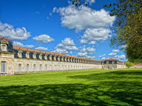 Long bâtiment historique en pierre Corderie Royale avec lucarnes et toit de tuiles orangées, devant pelouse verte et allée bordée d'arbres sous ciel bleu.