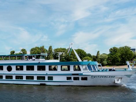 Bateau de croisière fluviale blanc MS Switzerland naviguant sur eau calme avec deux ponts de fenêtres, arbres verts et ciel bleu en arrière-plan.