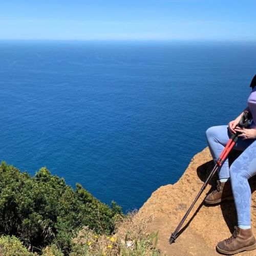 Randonneuse Elise assise au bord d'une falaise à Madère avec bâtons et sac à dos, contemplant l'océan Atlantique bleu et les falaises côtières.
