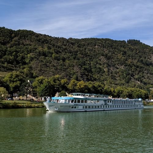 Bateau de croisière MS Switzerland naviguant sur eau verte calme avec collines boisées et petit village en arrière-plan sous ciel bleu.