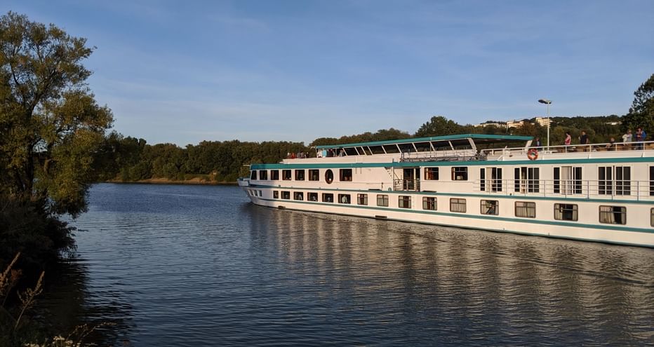 Bateau de croisière MS Switzerland naviguant sur un fleuve calme avec des arbres verts sur la rive gauche et des bâtiments visibles au loin.