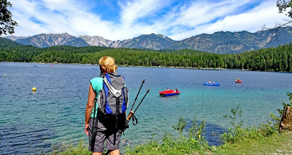 Randonneuse avec sac à dos et bâtons au bord du lac Eibsee, contemplant le lac turquoise avec pédalos et montagnes alpines.
